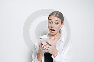 Surprised teenager girl with long hair in white shirt using mobile phone on white background