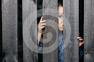 Surprised white boy looks out of the crack of a wooden fence. Childish curiosity. Espionage. Rural life. Child