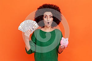 Surprised woman with Afro hairstyle in green sweater holding fan of dollars and piggy bank in hands