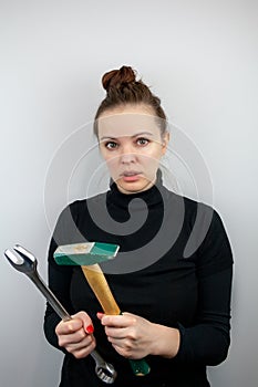 Surprised woman with bunched hair and black sweater holding a hammer and a big wrench in her hands