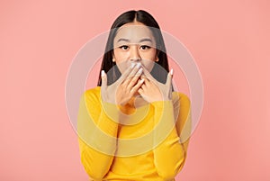 Surprised woman covering her mouth with hands while standing on pink background looking at the camera