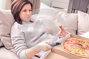 Surprised woman eating pizza and watching TV with remote control at home, warm tone