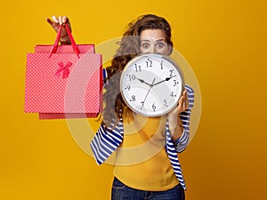 Surprised woman hiding behind clock and showing shopping bags