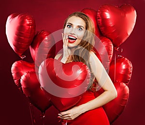 Surprised woman holding balloons red heart, portrait. Girl with red lips makeup wearing red dress. Gift, sale and Valentine`s day