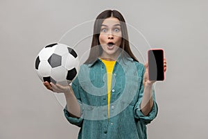 Surprised woman holding and showing blank display smartphone and football ball, betting on soccer.