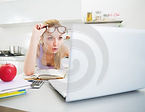 Surprised woman looking in laptop while studying in kitchen
