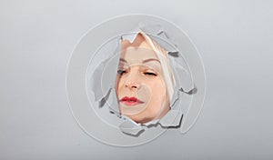 Surprised woman looking playfully in torn paper hole, has excited cheerful expression, looks through breakthrough of