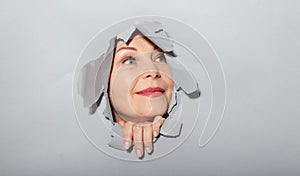 Surprised woman looking playfully in torn paper hole, has excited cheerful expression, looks through breakthrough of