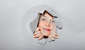 Surprised woman looking playfully in torn paper hole, has excited cheerful expression, looks through breakthrough of gray