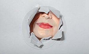 Surprised woman looking playfully in torn paper hole, has excited cheerful expression, looks through breakthrough of gray