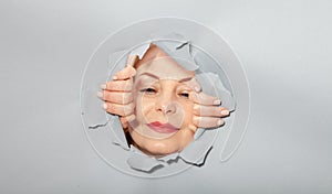 Surprised woman looking playfully in torn paper hole, has excited cheerful expression, looks through breakthrough of gray