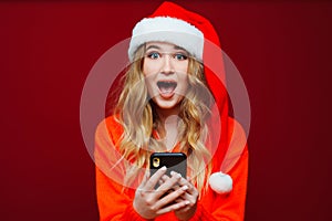 a surprised woman in a Santa Claus hat with smartphone on a red background.