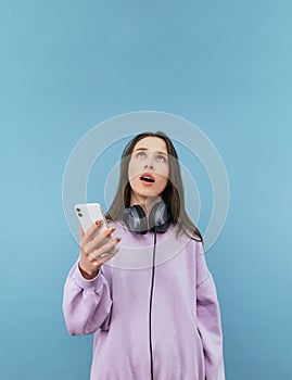 Surprised woman with smartphone in hands and headphones around her neck looking up at copy space with open mouth on blue