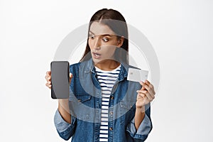 Surprised woman stare at her phone, showing smartphone screen and credit card, amazing prices online, shopping in