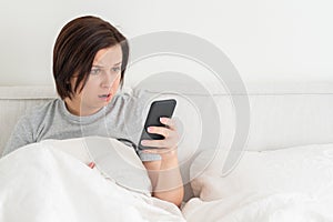 Surprised woman using smartphone as she sits in bed covered with duvet