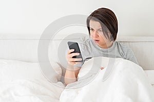 Surprised woman using smartphone as she sits in bed covered with duvet