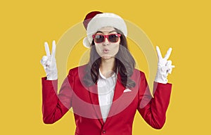 Surprised woman wearing in Santa style shows victory gesture with two hands on yellow background.