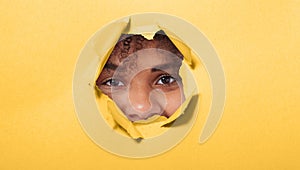 Surprised young African American woman looking playfully in torn paper hole, has excited cheerful expression, looks through