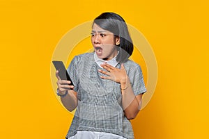 Surprised young Asian woman holding mobile phone with open mouth on yellow background