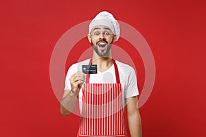 Surprised young bearded male chef cook or baker man in striped apron white t-shirt toque chefs hat isolated on red wall