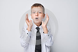 Surprised young boy in shirt and tie with hands near face against a gray background