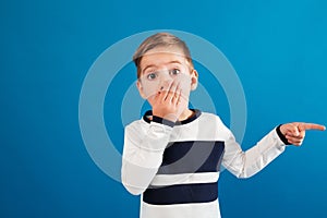 Surprised young boy in sweater pointing away and covering mouth