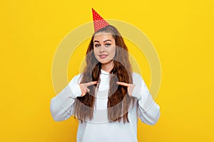 Surprised young brunette woman in birthday party hat points fingers at herself and looking impressed with disbelief, wearing white