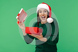 Surprised young brunette woman wear santa claus hat and green pullover opening gift box over green background.