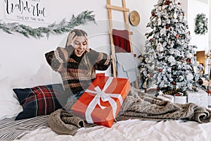 Surprised young caucasian woman with big Christmas present while sitting on a bed at home near Christmas tree