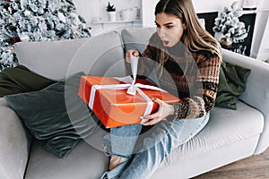 Surprised young caucasian woman holding big Christmas present while sitting on a sofa at home