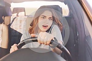 Surprised young cute female driver has telephone conversation via cell phone, drives car, looks with shocked expression as forgets