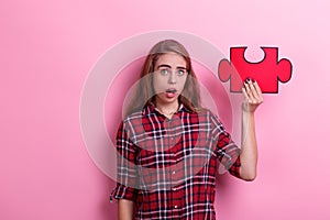 Surprised young girl, holds a large puzzle on her raised hand. On a pink background.