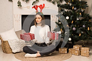 A surprised young girl sits and holds two red Christmas gifts near the white fireplace