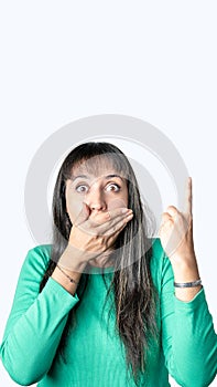 Surprised young Latin woman covering her mouth with one hand and pointing above with the other one