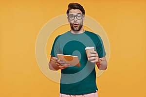 Surprised young man in casual clothing carrying digital tablet and cup