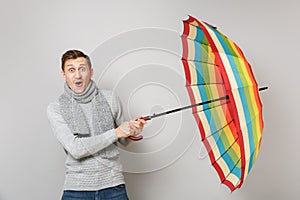 Surprised young man in gray sweater, scarf keeping mouth wide open, holding colorful umbrella on grey wall