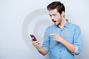 Surprised young man is holding phone and reading message pointing on it with finger