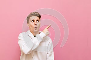 Surprised young man isolated on pink background and looks away and shows his finger on copy space. Guy in a white hoodie with