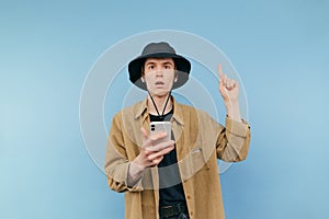 Surprised young man in panama and shirt stands on blue background with smartphone in hand and shows finger up with shocked face