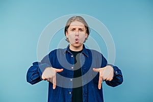 Surprised young man in a shirt stands on a blue background and shows his hands down on the copy space, looking at the camera with