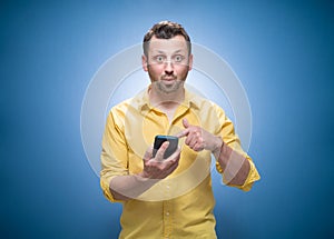 Surprised young man shows the mobile phone blank display - space for your text or image over blue background, dresses in yellow