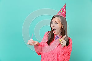 Surprised young woman in birthday hat with playing pipe holding cake with candle keeping mouth open pointing finger on