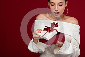 Surprised young woman with dark hair unpacking a red gift box, looking inside