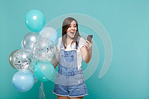 Surprised young woman in denim clothes looking on mobile phone celebrating, holding colorful air balloons isolated on