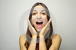 Surprised young woman with hands near open mouth looking at camera on gray background