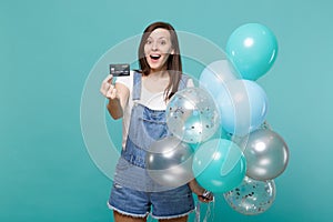 Surprised young woman keeping mouth open, holding credit bank card, celebrating with colorful air balloons isolated on