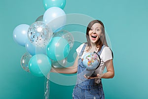 Surprised young woman keeping mouth wide open, holding Earth world globe, celebrating with colorful air balloons