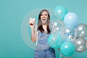 Surprised young woman keeping mouth wide open, holding piggy money bank, celebrating with colorful air balloons isolated