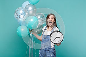 Surprised young woman keeping mouth wide open, holding round clock, celebrating with colorful air balloons isolated on