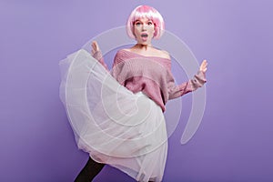 Surprised young woman in lush white skirt posing on purple background. Studio shot of fashionable female model in pink